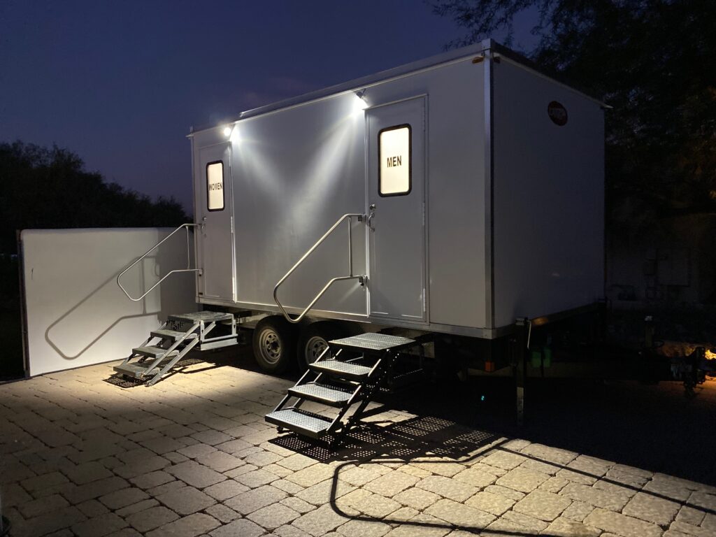 Luxury bathroom trailer at night with lit women's and men's doors over a stone patio, The Lavatory Los Angeles