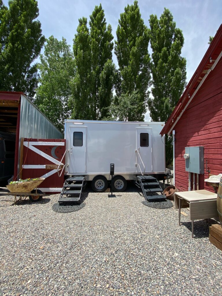 Bathroom trailer on gravel between two red barns with tall poplar trees, The Lavatory Los Angeles