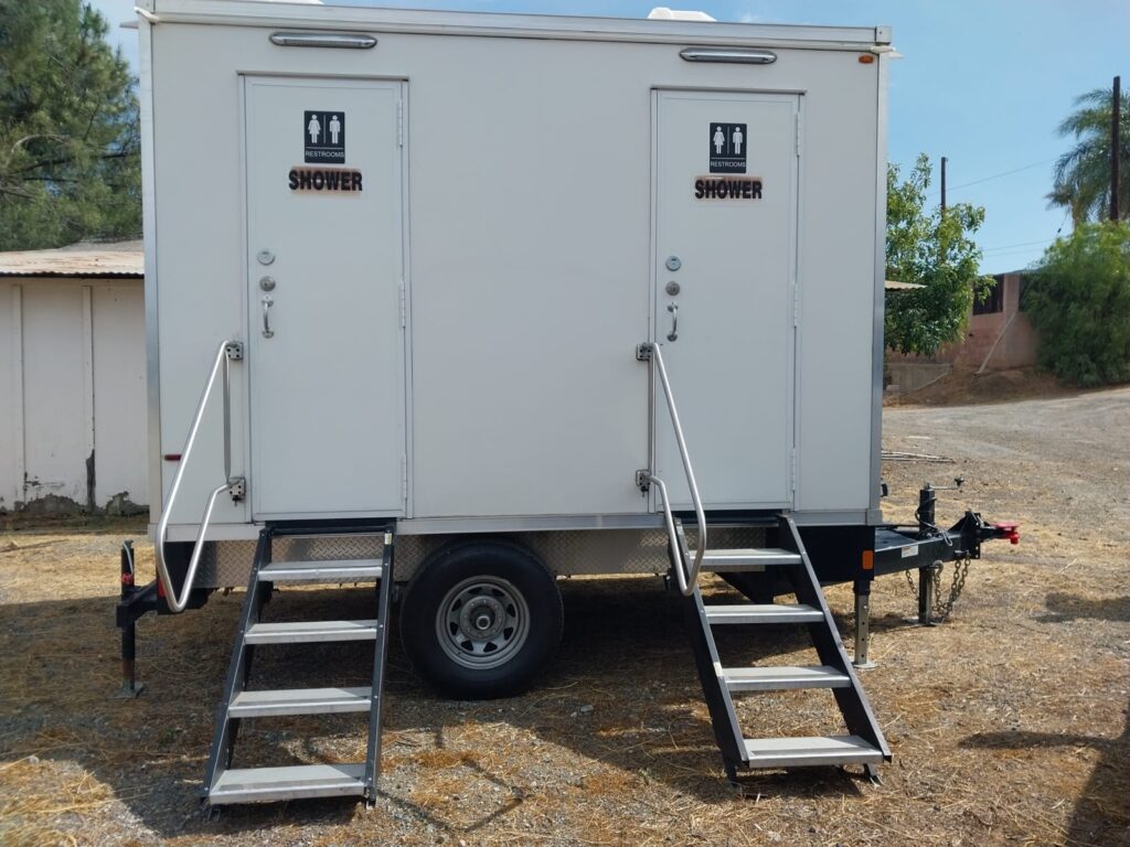 Front of a 2-station combo trailer showing both private shower room doors and stair rails on a dirt lot, The Lavatory Los Angeles