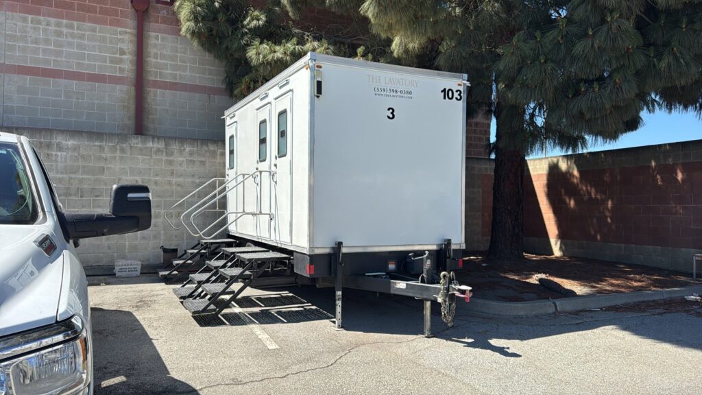 3-station private bathroom trailer parked in a lot beside a brick wall and a pickup truck, The Lavatory Los Angeles