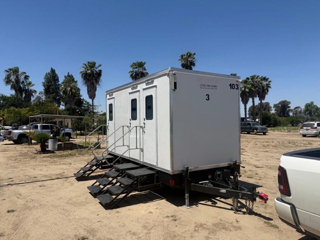3-station portable bathroom trailer set up on a dirt lot under tall palm trees, The Lavatory Los Angeles