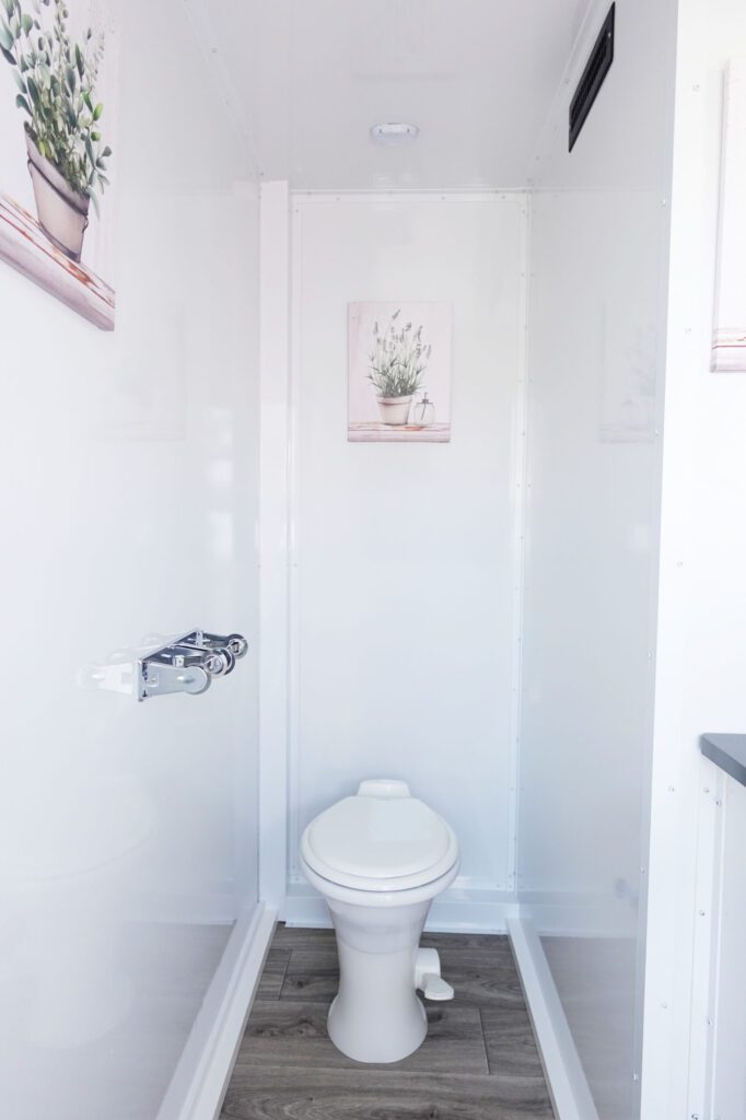 Bright white toilet room with wood-look flooring inside a 3-station portable bathroom trailer, The Lavatory Los Angeles