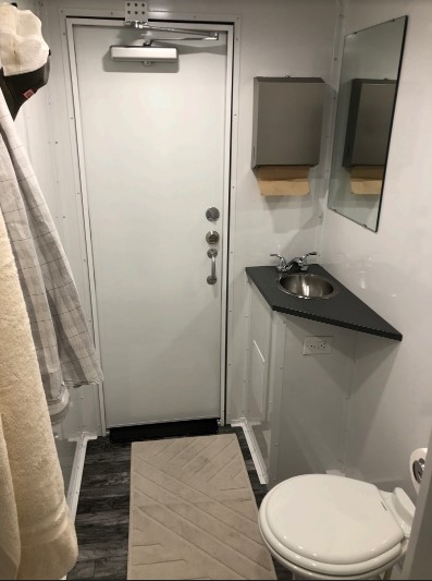 Private room inside a 4-station combo trailer with a white toilet, corner sink, paper towel holder and mirror, The Lavatory Los Angeles