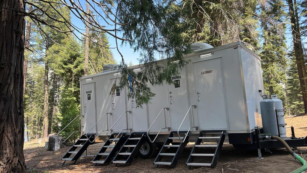 4-station combo trailer with shower and laundry doors set up under tall pines at a forest camp, The Lavatory Los Angeles