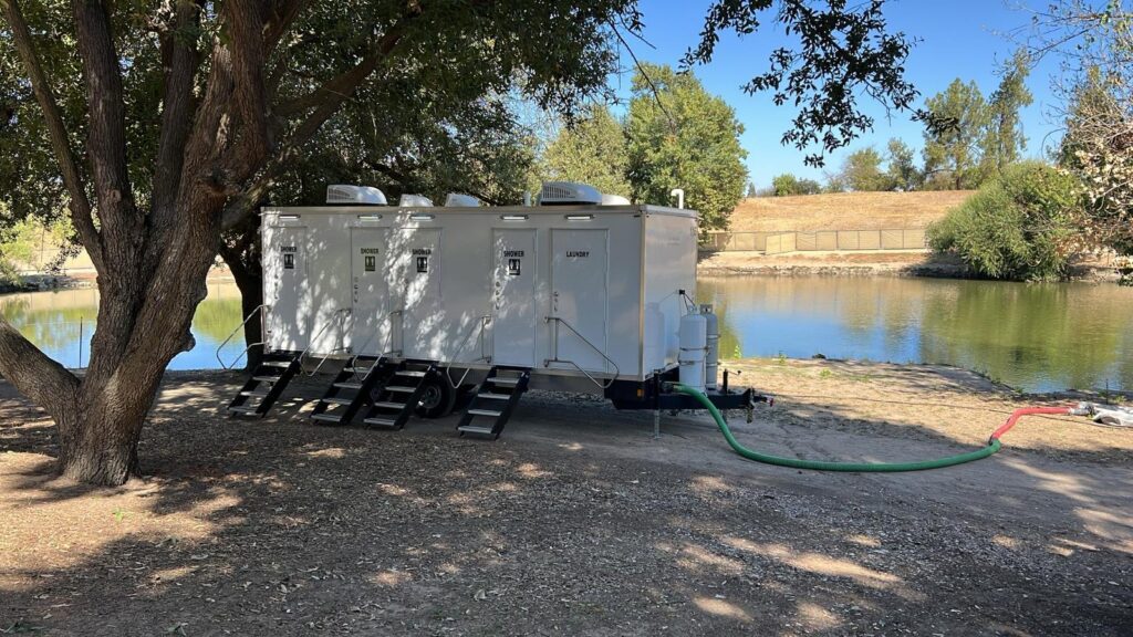 4-station combo trailer with four private bathroom doors and a laundry door, steps at each entry, parked under a shade tree beside a park lake, The Lavatory Los Angeles