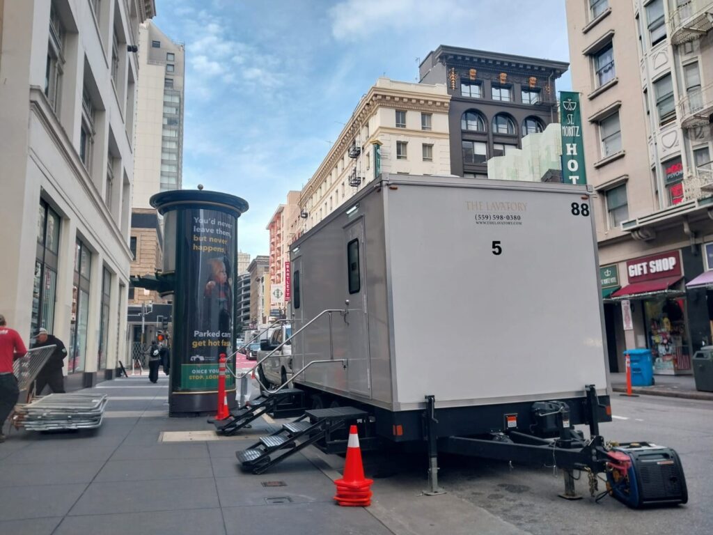 5-station temporary bathroom trailer at the curb on a downtown street while an event crew stacks barricades, The Lavatory Los Angeles