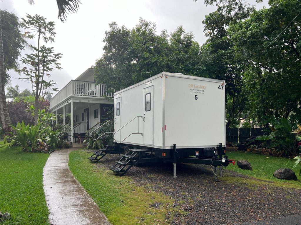 Luxury 5-station bathroom trailer parked on a gravel pad beside a garden path and a two-story home, The Lavatory Los Angeles