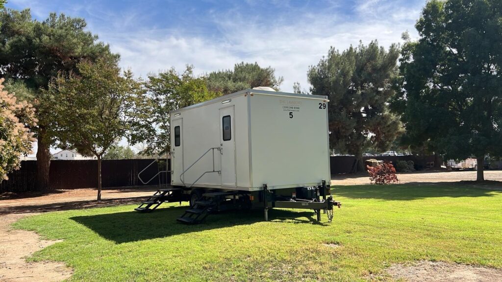 5-station mobile bathroom trailer on a sunny park lawn with tall pine trees, The Lavatory Los Angeles