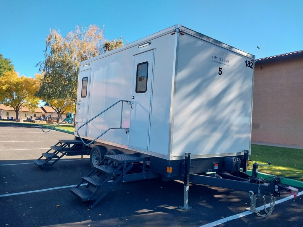 5-station portable bathroom trailer with two sets of steps in a school parking lot beside a playground, The Lavatory Los Angeles