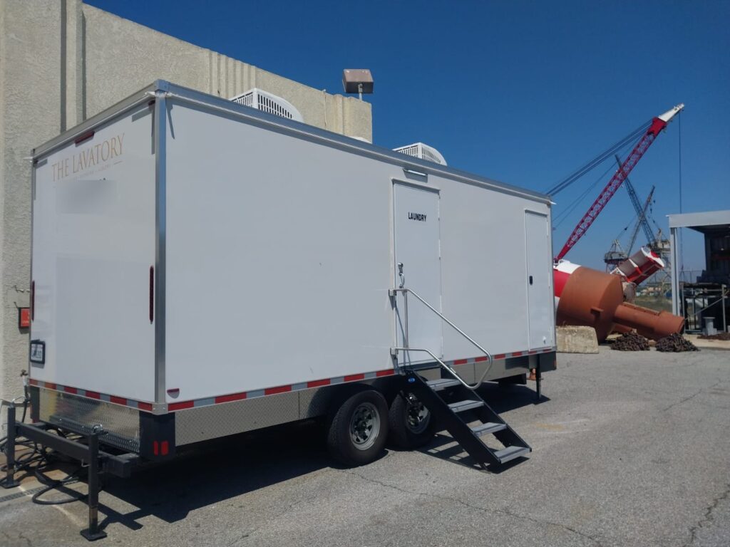 8-station laundry trailer with a laundry door and fold-down steps parked at a harbor yard beside a red crane, The Lavatory Los Angeles