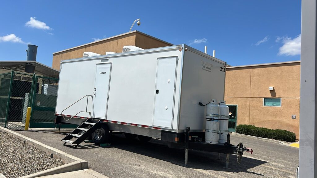 8-station mobile laundry trailer with a laundry door, fold-down steps and propane tanks parked beside a building, The Lavatory Los Angeles