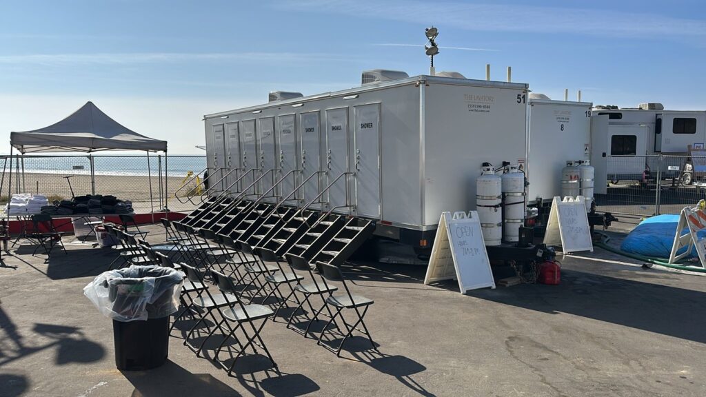 8-station shower trailer with folding chairs, a towel table and open-hours signs on a beachfront lot, The Lavatory Los Angeles