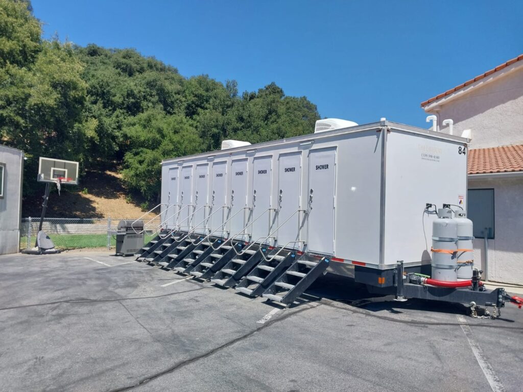 8-station shower trailer on a paved lot beside a basketball hoop and a grill below an oak-covered hillside, The Lavatory Los Angeles