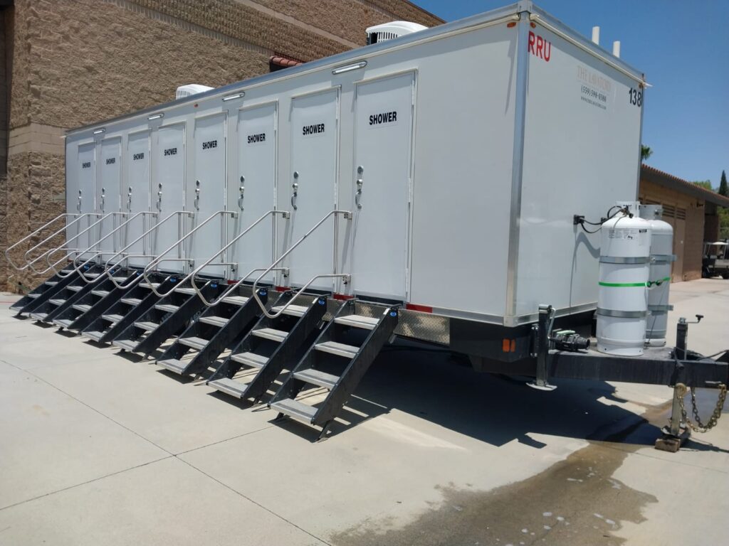 8-station shower trailer with fold-down steps and handrails at all eight shower doors, parked on concrete beside a block building, The Lavatory Los Angeles