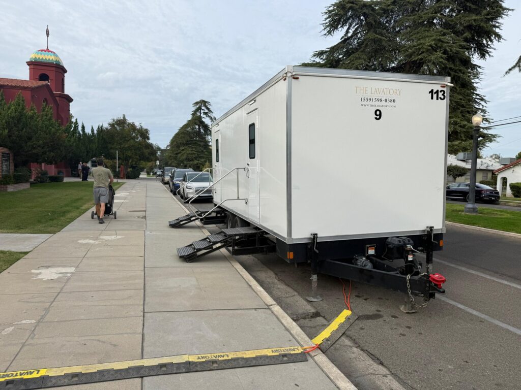 9-station portable bathroom trailer at the curb with steps on the sidewalk and a cable ramp as a man pushes a stroller past, The Lavatory Los Angeles