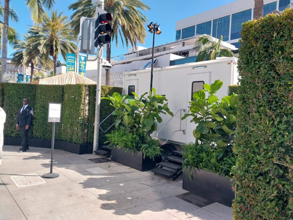 Hedge-screened 9-station luxury bathroom trailer with planters, a restroom sign and an event staff member in a dark suit on a palm-lined city street, The Lavatory Los Angeles