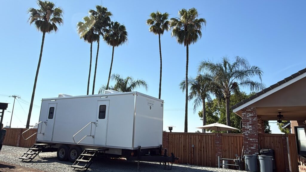 9-station portable bathroom trailer on gravel beside a backyard patio under tall palm trees, The Lavatory Los Angeles