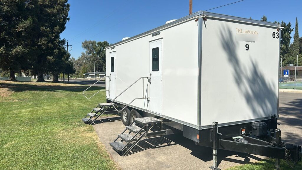 9-station bathroom trailer with two sets of steps and handrails parked on a park lawn beside tall pines, The Lavatory Los Angeles