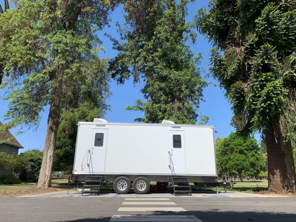 9-station luxury bathroom trailer parked under tall trees beside a park crosswalk, The Lavatory Los Angeles