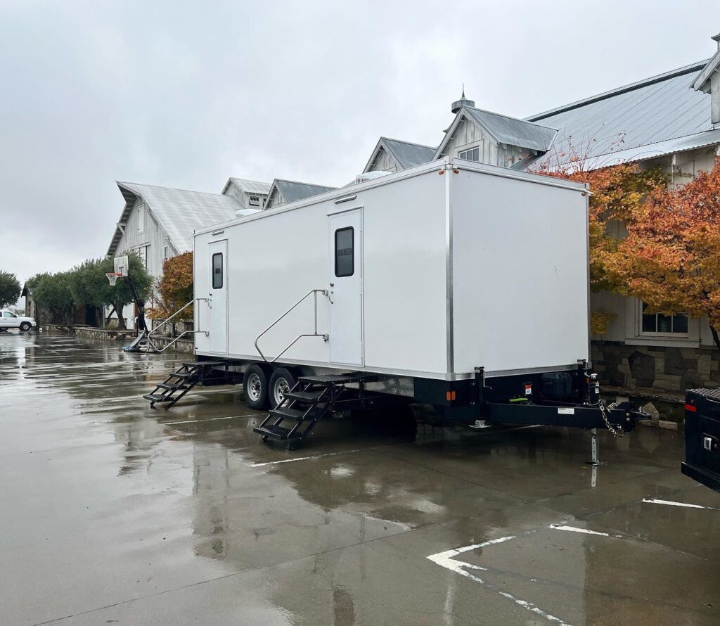 Portable bathroom trailer on a wet parking lot beside a large white barn event venue, The Lavatory Los Angeles