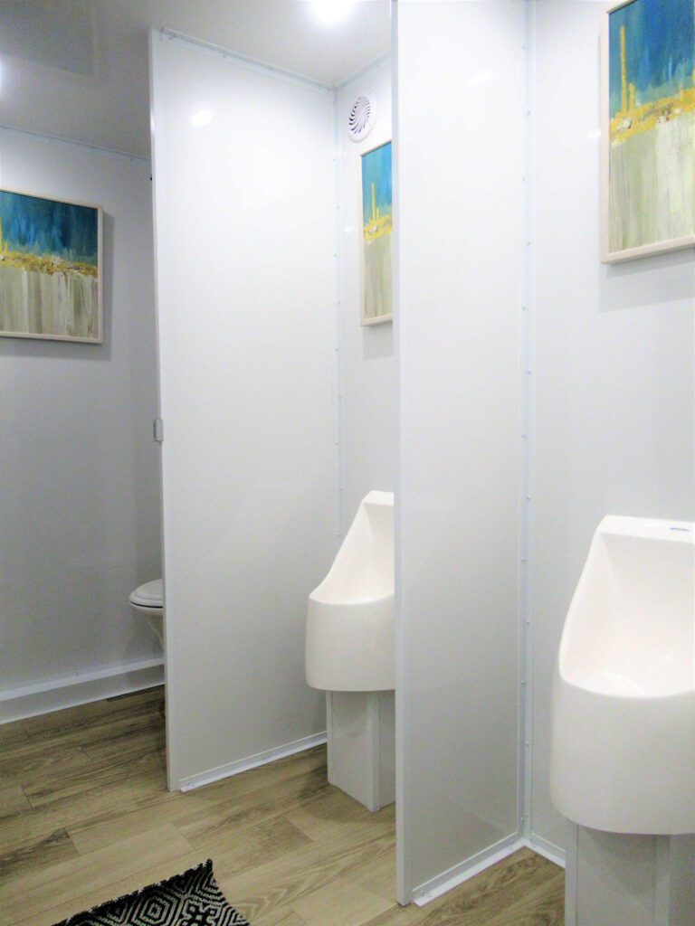 Two white wall urinals with privacy partitions and framed art on the men's side of a 9-station bathroom trailer, The Lavatory Los Angeles