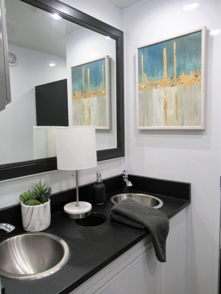 Vanity with steel sinks, black countertop, table lamp and framed art inside a 9-station bathroom trailer, The Lavatory Los Angeles