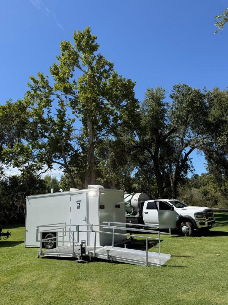 Accessible combo trailer with a ramp on a park lawn under tall trees, with a water service truck from our team behind it, The Lavatory Los Angeles