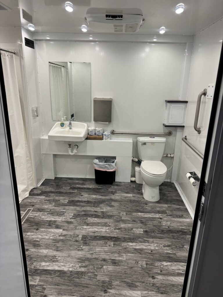 Inside an accessible combo trailer room with a wall-mounted sink, a toilet with grab bars, a wood-look floor and the roll-in shower curtain, The Lavatory Los Angeles