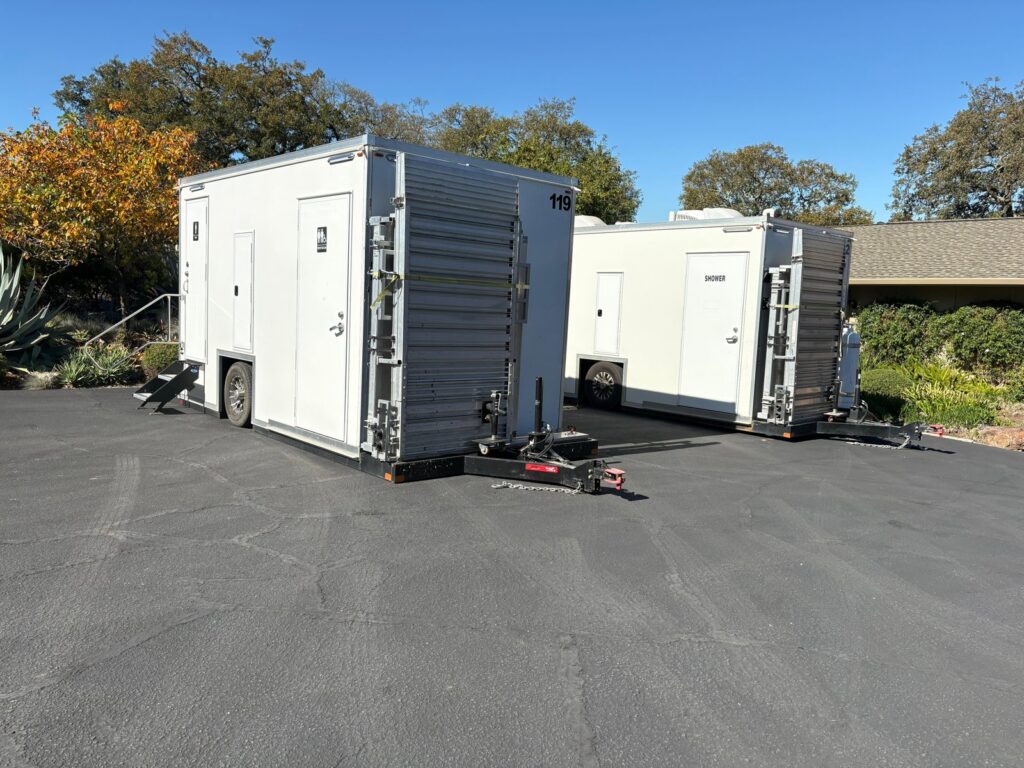Two accessible combo trailers with their ramps folded up for transport on a paved driveway, The Lavatory Los Angeles