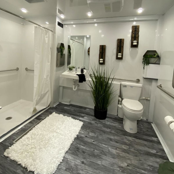 Full view of an accessible combo trailer room with the roll-in shower, a sink, a toilet with grab bars and a white bath rug, The Lavatory Los Angeles