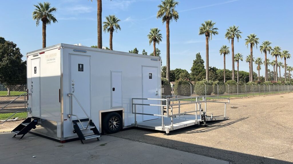 ADA plus 2 bathroom trailer with women's and men's doors, entry steps and a railed ramp landing under tall palm trees, The Lavatory Los Angeles