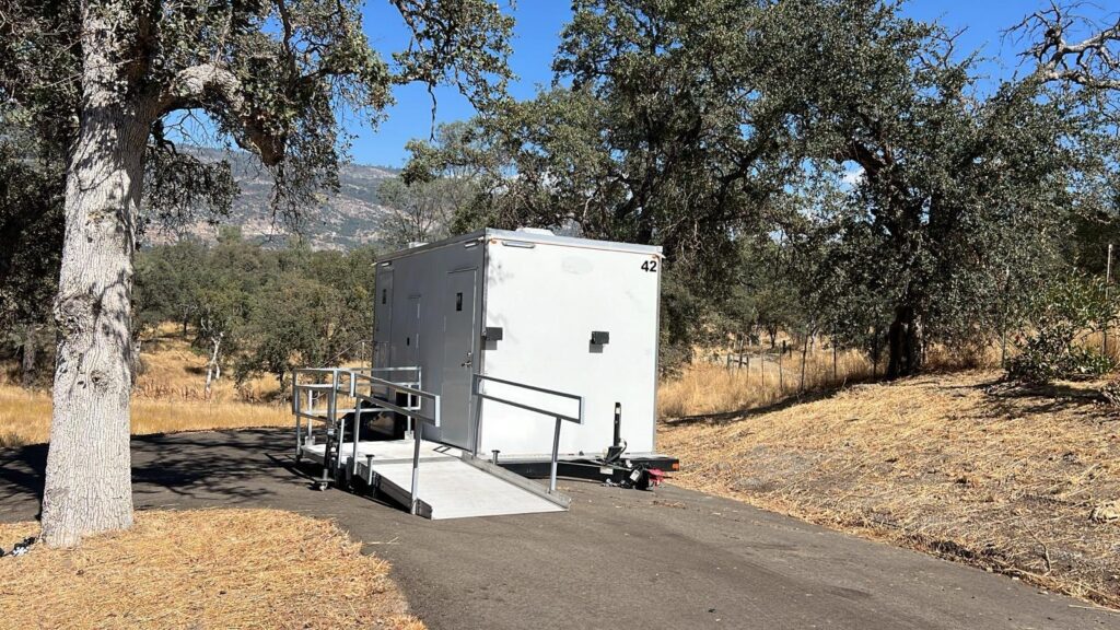 ADA plus 2 bathroom trailer with a railed wheelchair ramp set on a paved drive under oak trees at a rural venue, The Lavatory Los Angeles