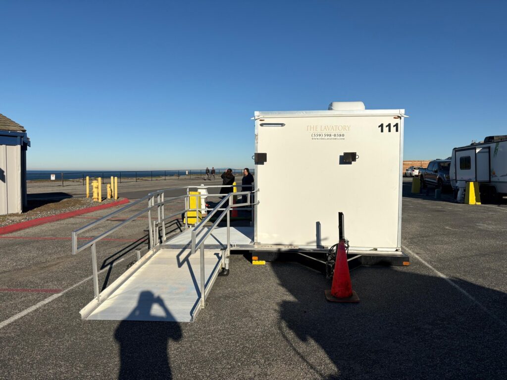 Two team members standing by the platform of an ADA plus 2 shower trailer with its ramp out in a beachside parking lot, The Lavatory Los Angeles