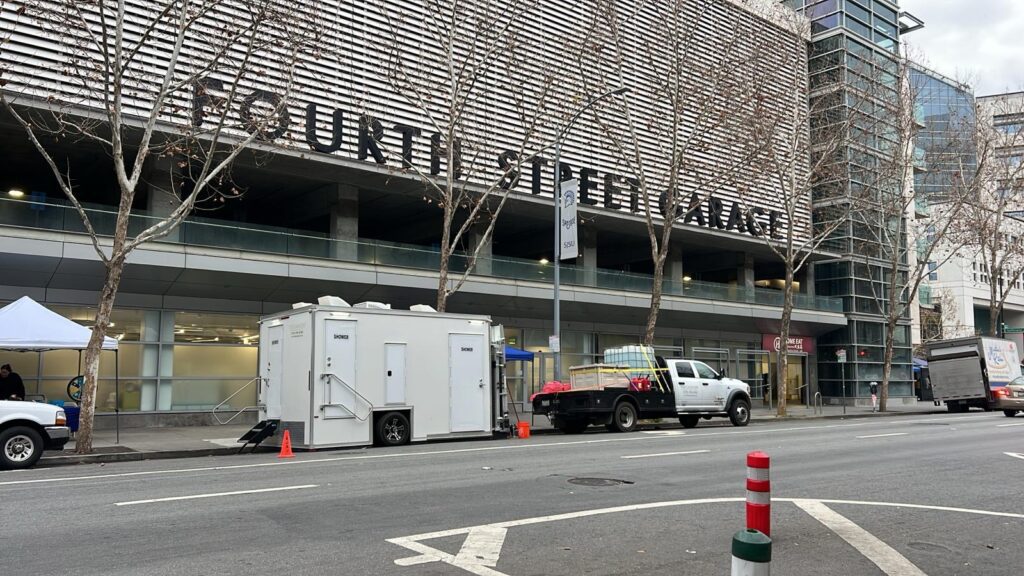Shower trailer parked at the curb on a downtown street beside a parking garage, with a vendor tent and a person on the sidewalk, The Lavatory Los Angeles