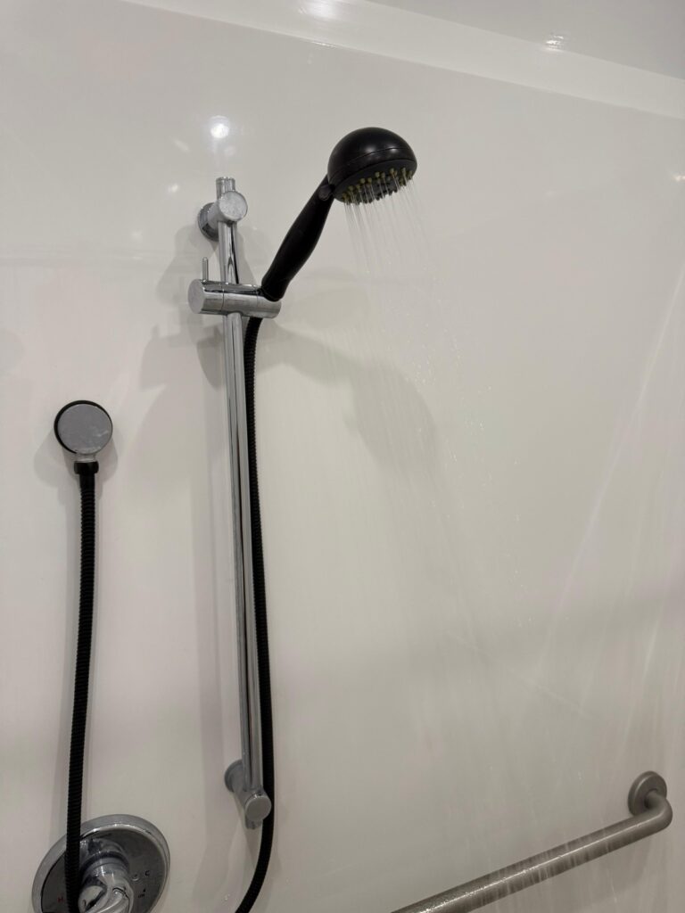Handheld shower head on a slide bar running hot water beside a stainless grab bar inside a shower trailer, The Lavatory Los Angeles