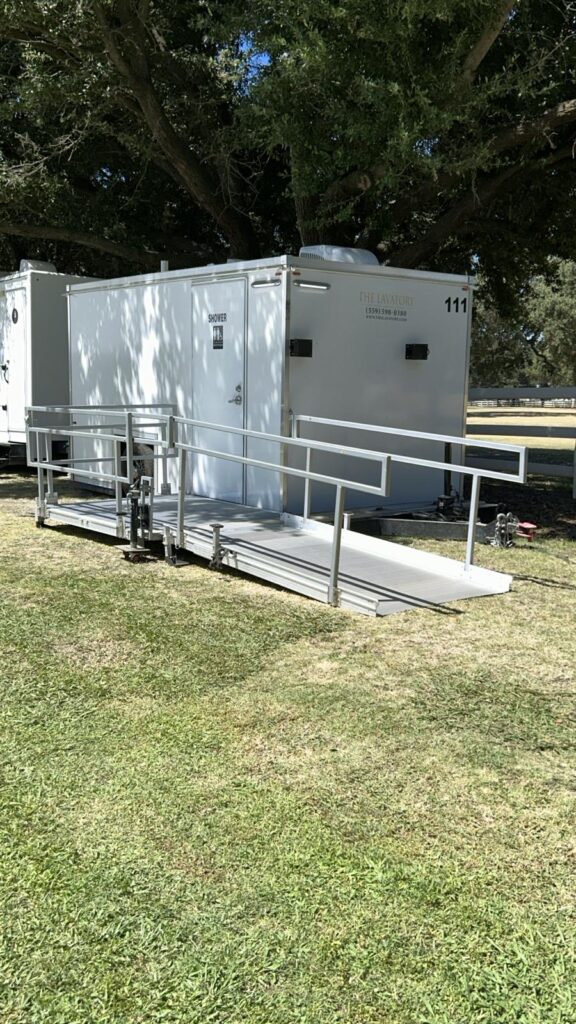 ADA plus 2 shower trailer with a railed ramp on green grass beside a second trailer under shade trees, The Lavatory Los Angeles