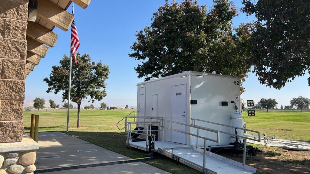 ADA plus 2 shower trailer with a railed ramp beside a park building and flag pole on a sunny day, The Lavatory Los Angeles