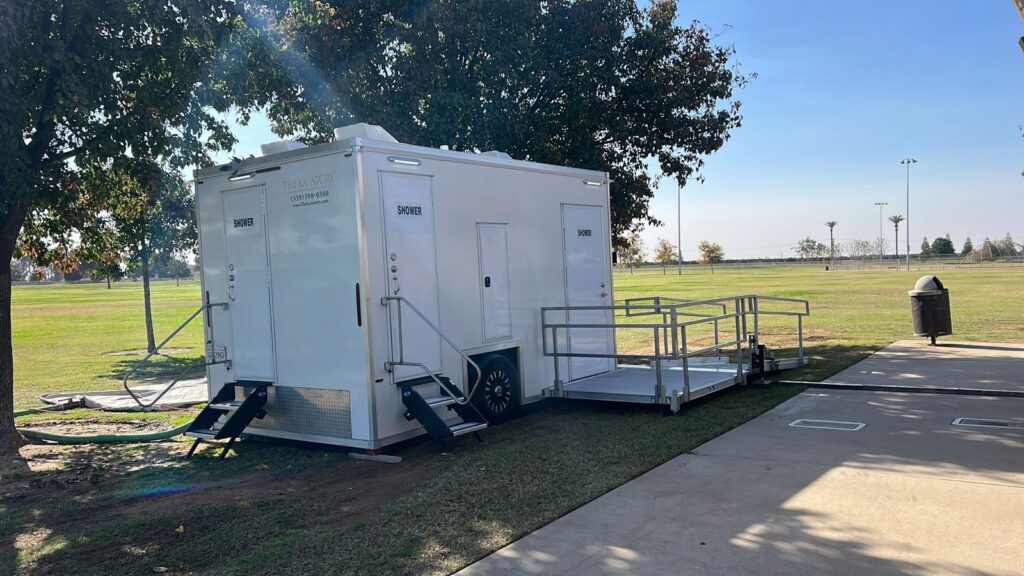 Private shower trailer with two stepped shower doors and a railed accessible platform under shade trees in a city park, The Lavatory Los Angeles