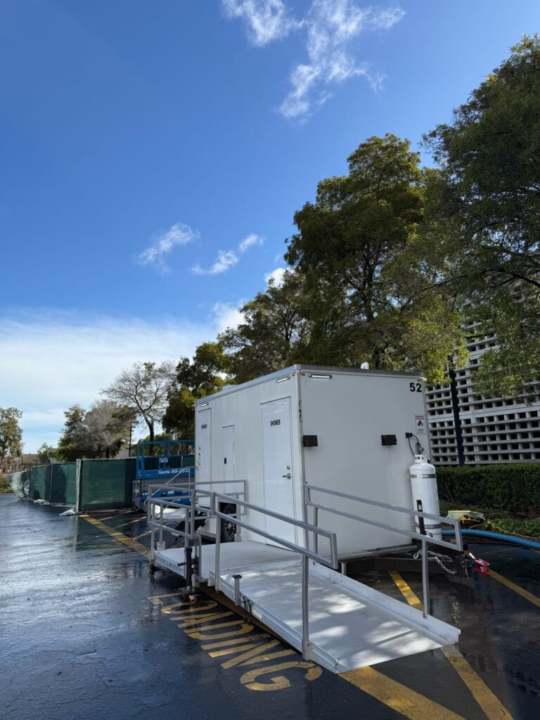 ADA plus 2 shower trailer with its ramp down on a wet parking lot beside tall trees after rain, The Lavatory Los Angeles