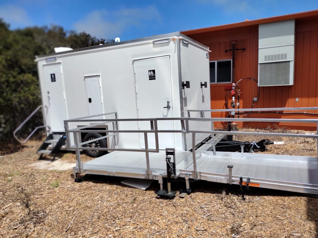 ADA plus 2 bathroom trailer with a ramp with handrails set on wood chips beside a building, The Lavatory Los Angeles