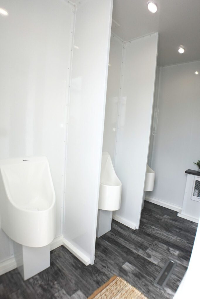 Row of white urinals with privacy partitions in the men's room of an ADA plus 6 portable bathroom trailer, The Lavatory Los Angeles