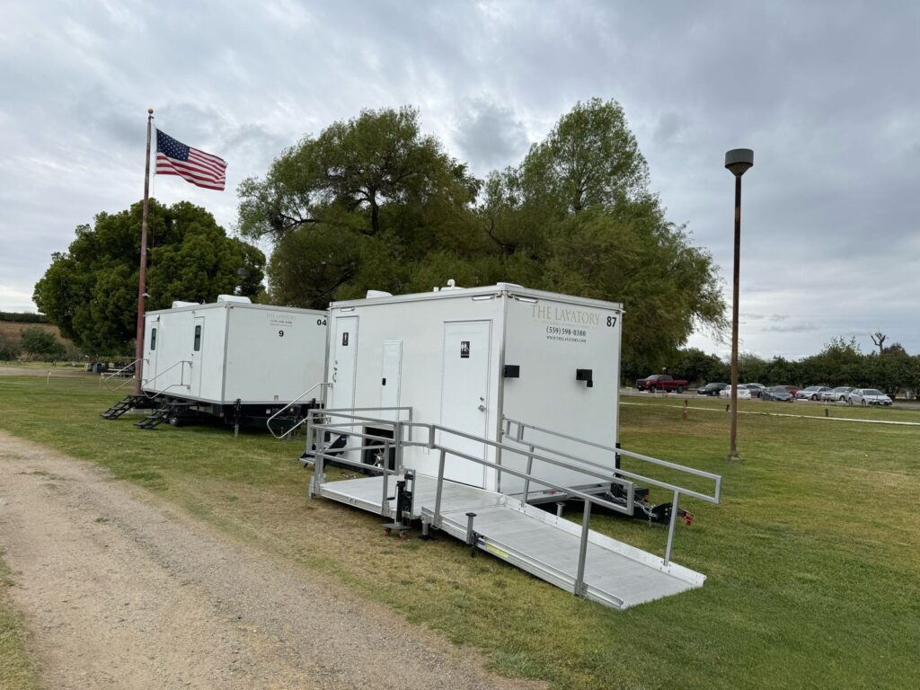 9-station bathroom trailer and an accessible trailer with a railed ramp set on a park lawn by a flagpole, The Lavatory Los Angeles