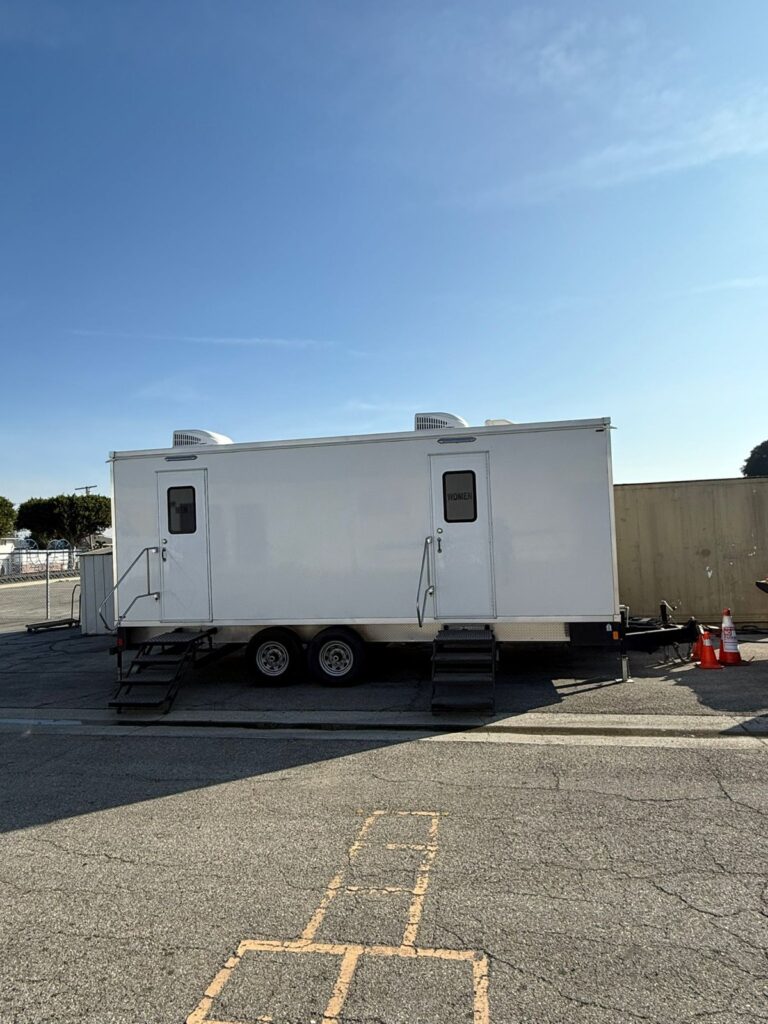 Long bathroom trailer with two doors and steps parked on a school blacktop next to a painted hopscotch court, The Lavatory Los Angeles