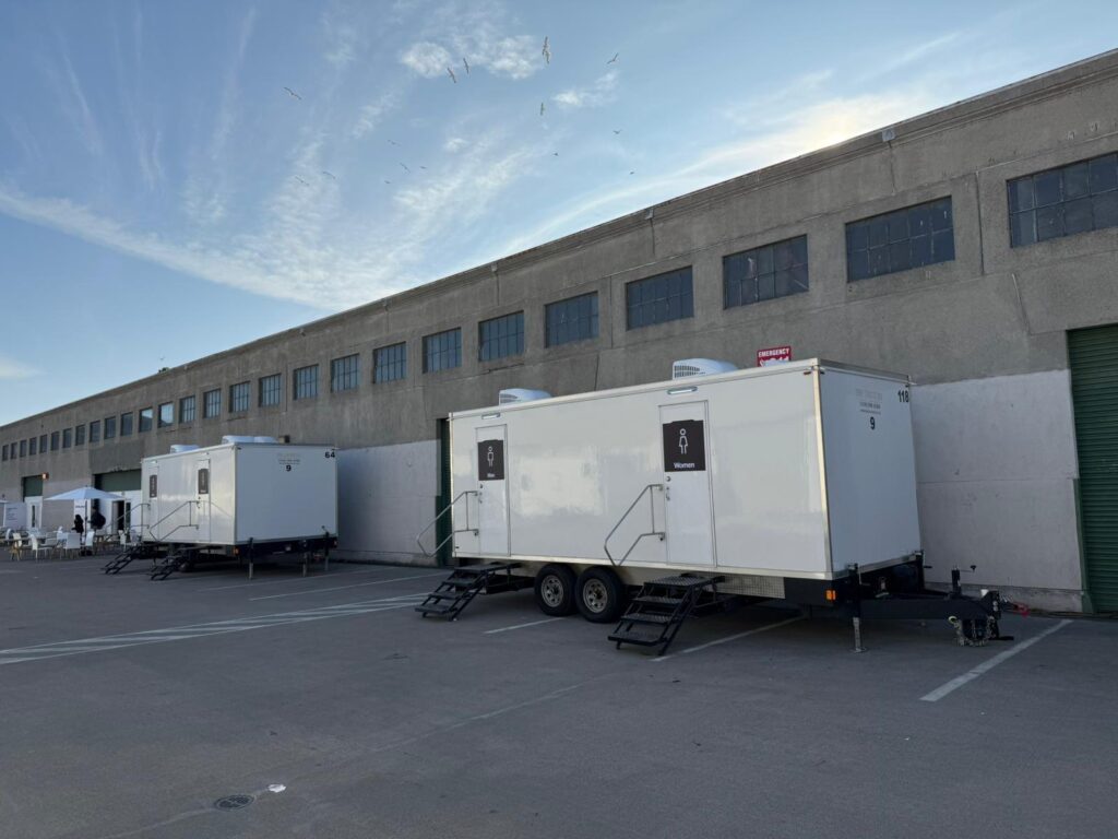 Two bathroom trailers with steps lined up along a long concrete warehouse for an indoor company event, The Lavatory Los Angeles