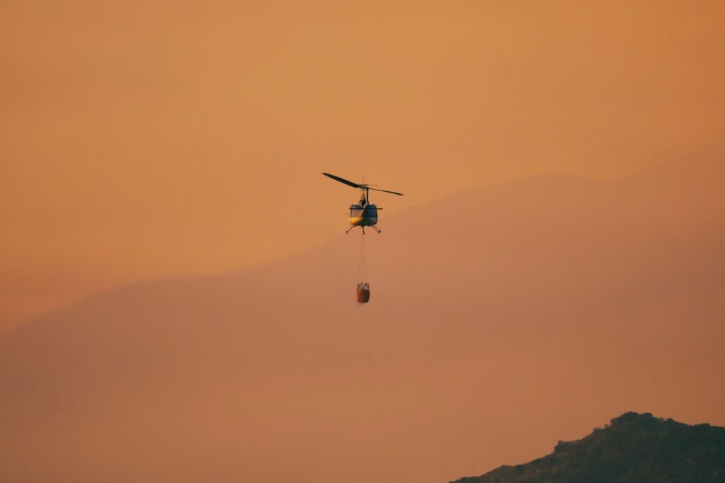 Firefighting helicopter carrying a water bucket through orange smoke over Southern California hills, The Lavatory Los Angeles