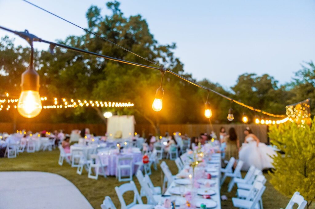 Backyard quinceanera reception at dusk with long tables, string lights and guests near the guest of honor, The Lavatory Los Angeles