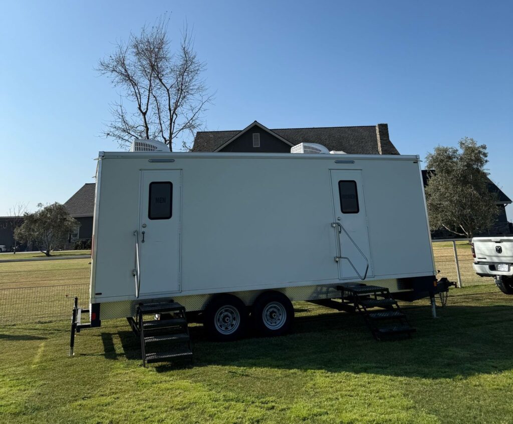Long portable bathroom trailer with two entry doors and steps set on the back lawn of a large country home, The Lavatory Los Angeles