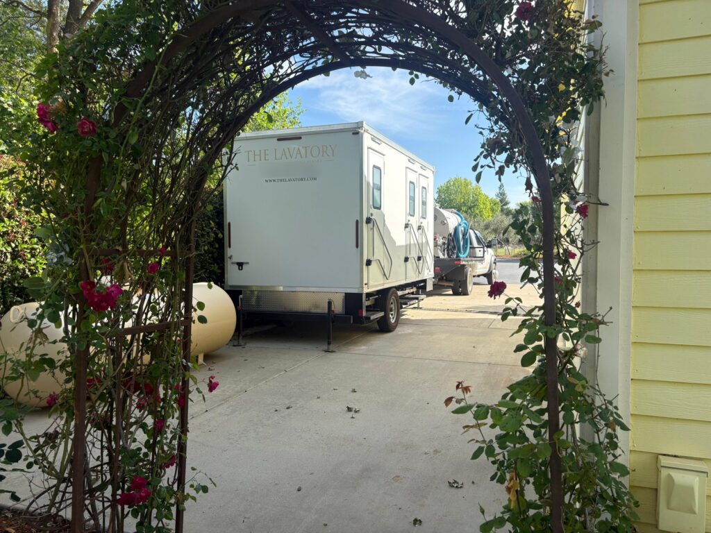 Bathroom trailer parked in an estate driveway, seen through a garden arch of red roses, with our service team's pumping truck behind it, The Lavatory Los Angeles