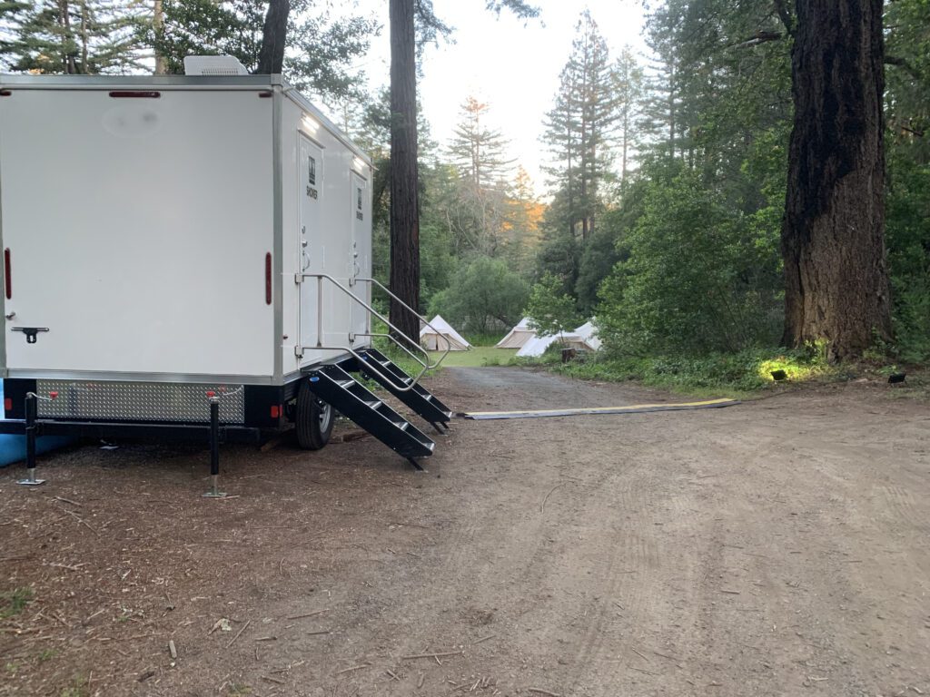Bathroom trailer parked on a forest dirt road near tents and tall trees, The Lavatory Los Angeles