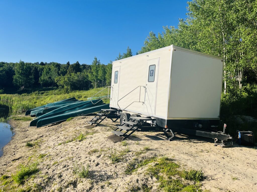 Bathroom trailer on a sandy lakeshore next to stacked green canoes, The Lavatory Los Angeles